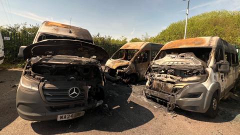 Three minibuses in a car park. They are all clearly destroyed by a fire, showing melted plastic, no glass in the windows etc.