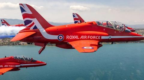 A close-up aerial image of Red Arrows aircraft, flying over the blue sea in close formation. One has red, white and blue smoke emerging from the back.