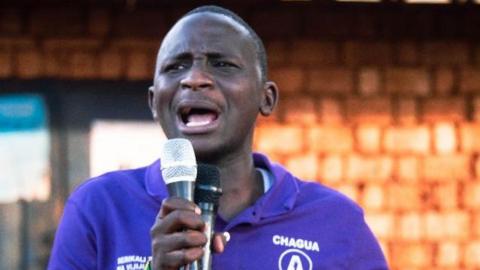 Abdul Nondo in a purple shirt speaking into a microphone at a campaign rally.