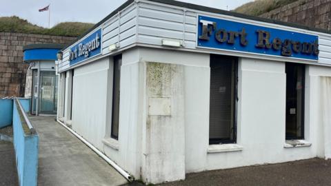 A single storey white building with blue signs with sliver letters reading Fort Regent on both sides. The building has windows with closed blinds and has signs of damp. There is a ramp with a blue hand rail leading to a rounded entrance with glass doors. Surrounding the building is a brick wall behind which is two hills which have a UK flag flying between them.