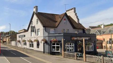 A Google street view image of the Whyteleafe Tavern, which is grey and has hanging baskets of flowers over the windows. 