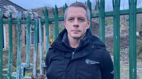 A man wearing an Environment Agency jacket, standing in front of a green bolted gate - behind that is a pile of waste that was dumped there illegally.