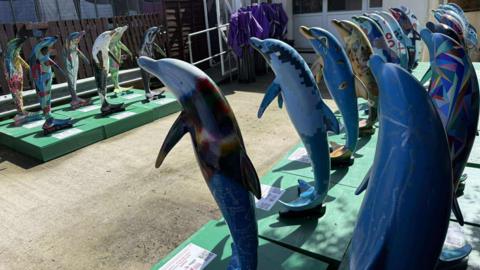 Double rows of the dolphin sculptures mounted on green blocks face each other in a concrete yard. They sit upright on their tails and every one is uniquely decorated. They are brightly coloured and individually patterned. 