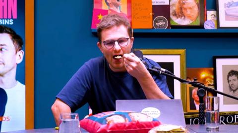 A man with a blue t-shirt and glasses, holding a spoon going into his mouth, in a podcast studio.