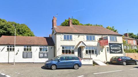 The exterior of a pub with white walls and a black sign on it saying 'The Old Stag's Head, Banks's Ales & Spirits'. A red for sale sign is attached to the building. Two cars are parked outside the building.