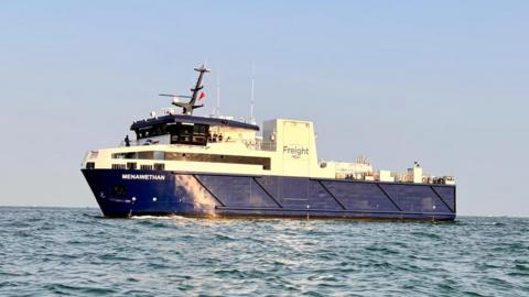 The new freight vessel out at sea. It is blue at the bottom and white at the top with the name of it clearly visible. The sun is shining down on it.