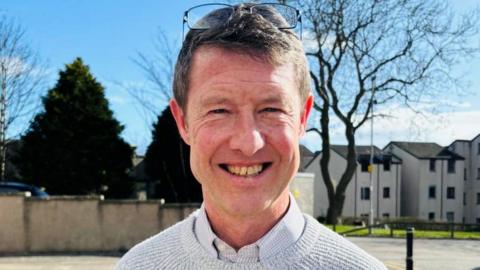 Ian Rodger, a man looking at the camera and smiling. His glasses are on his head and he is wearing a light checked shirt under a light great sweater. There are houses behind him, and trees.