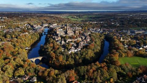 An aerial photo of Durham City with the Cathedral and other buildings visible on land surrounded by a river. The leaves on the trees are turning orange and brown.