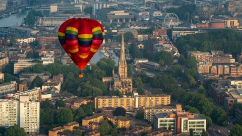 A multi-coloured, mainly red, hot air balloon flying over Bristol. Trees, buildings, and some of the city's landmarks can be seen beneath it including Bristol Cathedral, the harbourside and a ferris wheel.