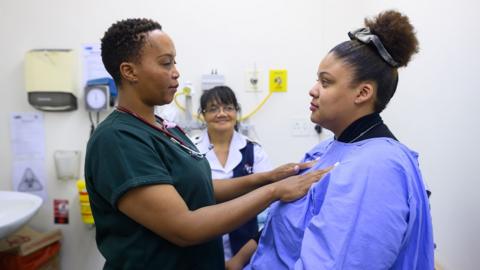 Stock photo shows a healthcare practitioner assessing a patient's breasts through a hospital gown as a healthcare assistant looks on in the background, in a clinical setting.