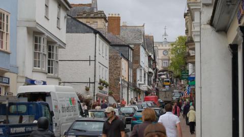 A busy shopping street in Totnes with cars and vans heading up the road under a cream arched gatehouse which has a clock face on it. There are lots of people walking up and down Fore Street.