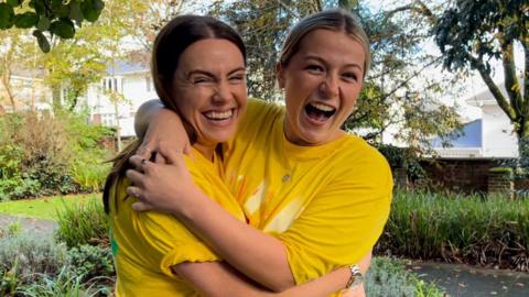 Two women - Lucy Bolt and Charlotte Cory - hug each other and laugh in a garden while wearing yellow Children in Need t-shirts.