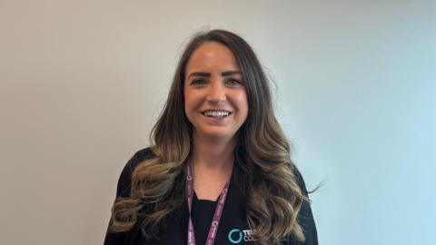 Jodie Allen has long brown hair and is wearing a black jacket with a black t-shirt that has the telford college branding on it. She is wearing a purple lanyard