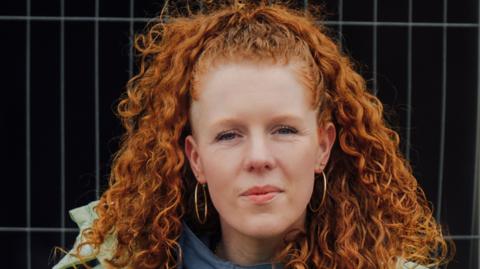 A woman with long curly red hair and large gold earrings standing against a dark background