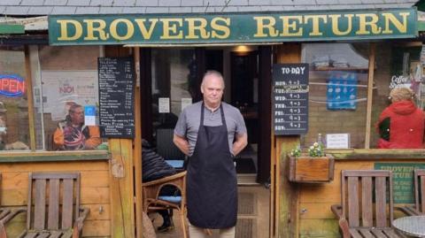A man, standing in the doorway of a cafe, wearing a black apron, and a grey top. People are inside the building. There are signs above and either side of the door and chairs and a table outside. The man looks serious. 