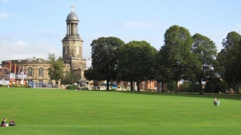A landscape photo showing a mother and child walking across a large patch of grassland. Two people are sat down to the left of the photograph. In the distance a line of trees leads up to a built up area with an historic looking building with white and black beams on the front of it. A very large building behind it has arched windows and a tall tower with a dome shape on the top. At the top is a crucifix. 