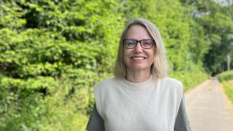 Jess Bailey, Devon County Council member for Ottery St Mary, in a lane next to the proposed quarry site, wearing a beige woollen tanktop and glasses.
