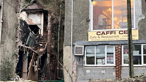 Collapsed building next to cafe and restaurant with sign: Caffi Ce and a telephone pole blocking the rest of the sign