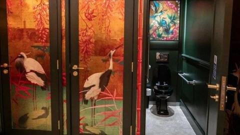 A black toilet on the right in a toilet cubical. On the left there is a red and yellow tropical design with flamingos on the cubical doors.