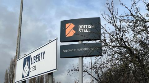 The signs at the front of the British Steel plant in Scunthorpe - one reading 'Liberty Steel' and another reading 'British Steel - building stronger futures'. They are partially obscured by trees.