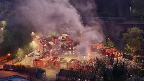 Drone footage of an industrial area of skips and shipping containers, with white smoke rising above