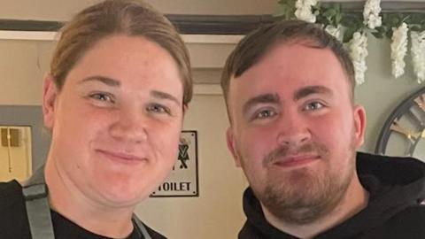 Luke Littler and a member of staff at the cafe. Zoe is wearing an apron and he is wearing a hoodie. They are both smiling. There is a toilet sign behind them. 