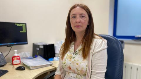 Dr Michelle Le Cheminant is wearing a white dress with patterned pale flowers and a creme cardigan. She has medium length light brown straight hair.