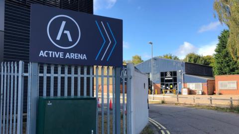 A sign of outside of Active Arena. It is an industrial estate with railings and corrugated deep blue buildings.