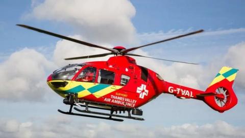 A red helicopter in flight with "SUPPORT THAMES VALLEY AIR AMBULANCE" written on the side
