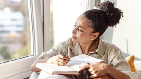 Someone looking thoughtfully out of a window while writing in a journal.