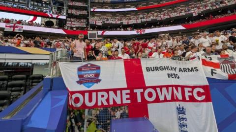 The giant white flag with a red St George's Cross with "Bourne Town FC" in white letters across the middle. It also has the club's badge and the England badge on it as well as "Burghley Arms, PE10" written in the top right. There are hundreds of England fans in the stadium behind it. To the right is another England flag, with the Grimsby Town logo on it.