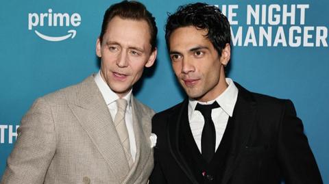 Tom Hiddleston stands on the left wearing a light suit. A man with dark hair and a dark jacket and tie stands to his left