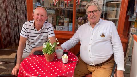 Two men smile while sitting at a small red circular table outside the front of a cafe. A pot plant is on the table along with cups of coffee. The store behind them has an orange frame with historic items in the window.
