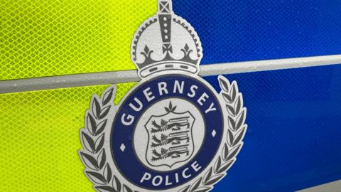 A coat of arms featuring three blue lions, with a laurel wreath and the words Guernsey Police  surrounding them is on a fluorescent yellow and dark blue background.