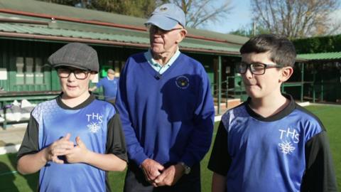 Two boys are wearing blue and black school PE tops as they stand on a bowling green with the single-storey club house behind them. They are standing either side of a man who is wearing glasses, a light blue baseball cap and a blue sweatshirt and trousers.