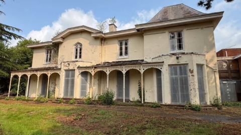 A large mansion style property, with peeling cream paint and shuttering over the large downstairs windows