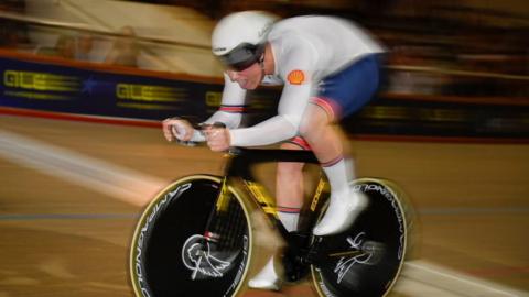 A young man in white and blue GB track cycling kit and white helmet racing on his black bike in a velodrome. The background is blurred with some spectators watching. 