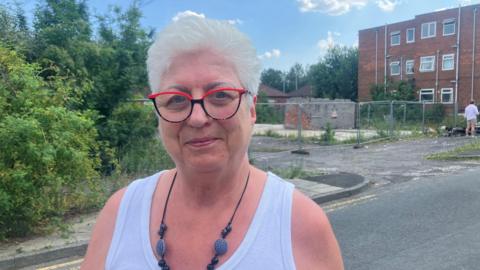Baguley Crescent resident Francesca Estasy is pictured in front of wasteland opposite her apartment. She has short, styled white hair and wears red and black rimmed glasses. She has a lilac vest top and a low necklace in black and blue. Behind her, security fencing runs between one three-storey apartment block and a piece of flat ground leading to overgrown bushes.