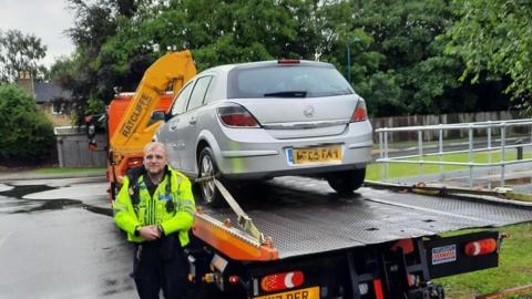 Officer standing in front of a seized car on a trailer 