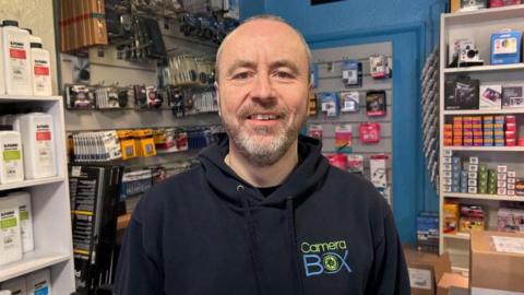 Adrian Clarke with very short dark hair and beard, wearing a Camera Box hoodie and standing in his shop. Shelves are visible with boxes of film, smart cards, cameras and accessories behind him.