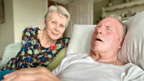 This is a head and shoulders picture of Anne Scowen and her husband Ray. Ray is lying on a bed with his head on a pillow in the foreground and has his eyes shut. Anne who has short grey hair and is wearing a floral dress, is sitting in a chair next to him, smiling.