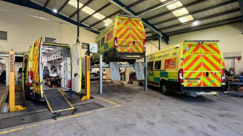 Three ambulances are being worked on inside a depot. Two are on hydraulics, with the middle ambulance lifted into the air. The rear doors are closed on the third ambulance. 