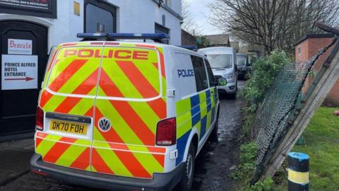 A police van is parked on a muddy track beside a white pub building and a wire fence which has been damaged and is leading to one side. In the background a large grey van is visible