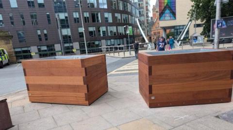 Two large brown planters on a pavement with a road behind with pedestrians walking across the road towards it. 