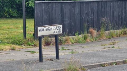 A road sign saying Waltham Close. It is on a pavement covered with weeds.