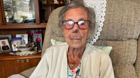 An elderly woman with short grey hair, glasses, a white cardigan and floral top, and a necklace, looks straight at the camera while sitting in a large armchair. Behind her is a wooden and glass cabinet with pictures.