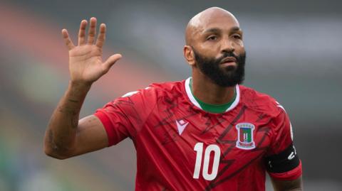Emilio Nsue, wearing a red Equatorial Guinea shirt with a number 10 on the chest, holds his right hand aloft during a football game. The player has a bushy black beard and a neutral expression on his face