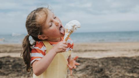 Little girl stands by the ocean on a beach and grips a wafer ice-cream cone. She licks around the vanilla ice-cream in an attempt to catch the drips.