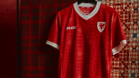 In the picture a red Wales football top with white trim can be seen. There is a logo that reads SUDU on the right and the Welsh flag with the red dragon on the left. The background is a patterned wall with a mixture of different designs and colours, including red, green and cream.