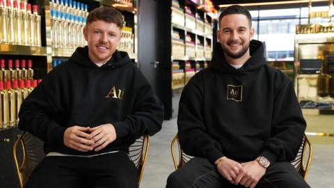 Two men in their twenties wearing black hoodies, featuring a gold Au logo, sit on gold chairs. Behind them the shelves are stacked high with gold vodka bottles.